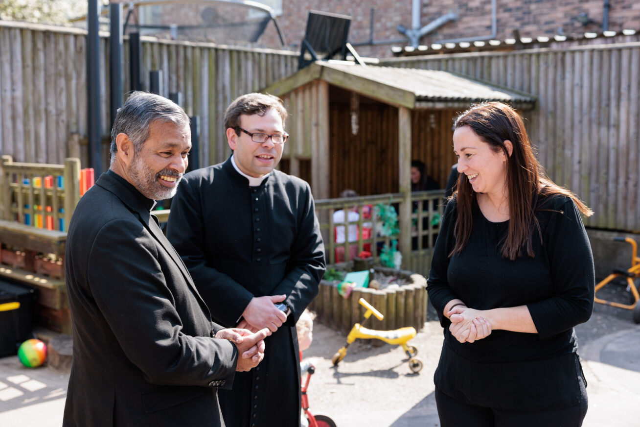 Bishop John talking to Father Daniel Howard and a female staff working at St Columbas nursery. The picture is outside with a blurred image of toddler in an outdoor play area wi
